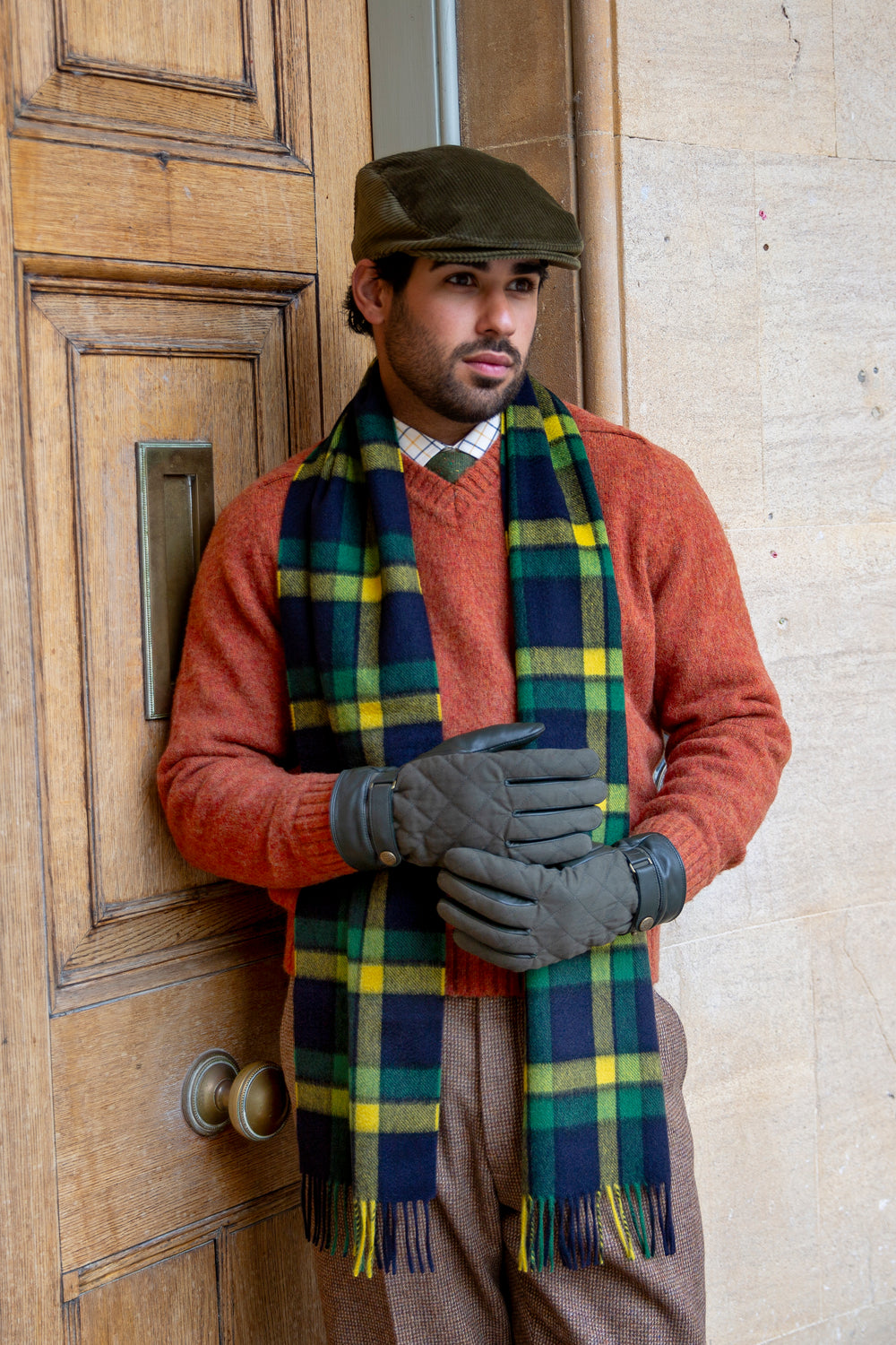 Man wearing a check cashmere scarf,  quilted gloves and a flat cap stood by a large door