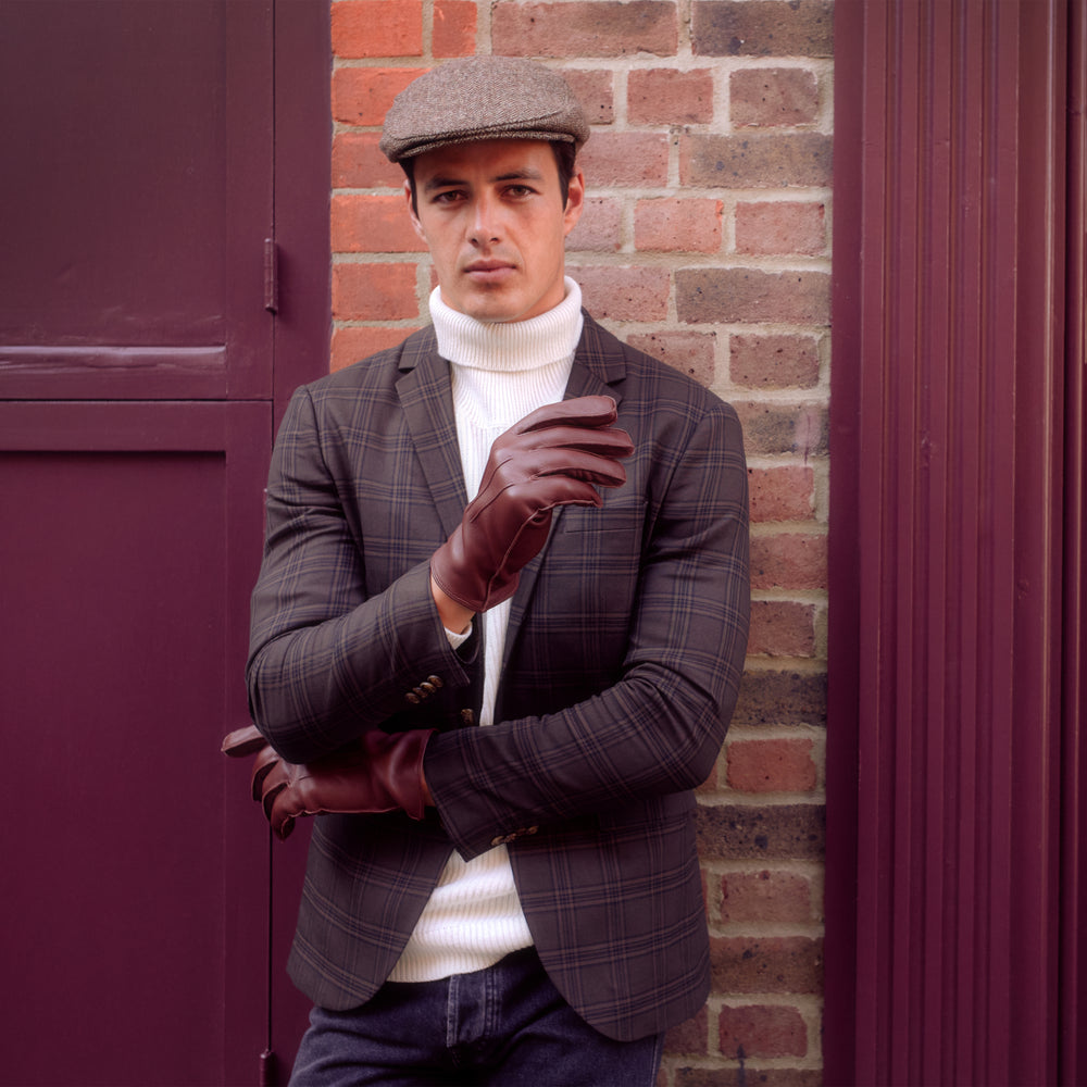 Man wearing a tweed flat cap and leather gloves by a brick wall