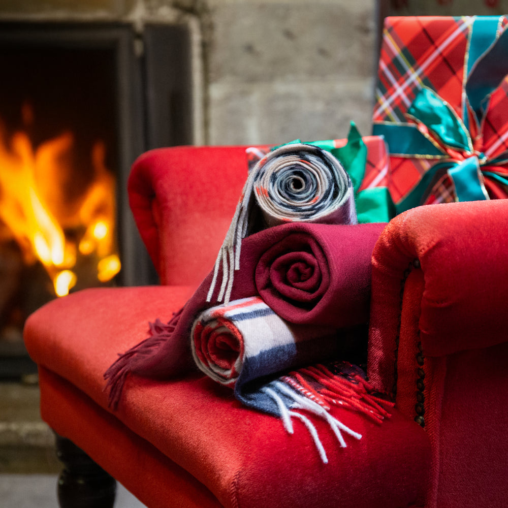 A cozy holiday scene with rolled plaid and plain cashmere scarves draped over a red armchair beside a fireplace, with wrapped Christmas gifts in the background.
