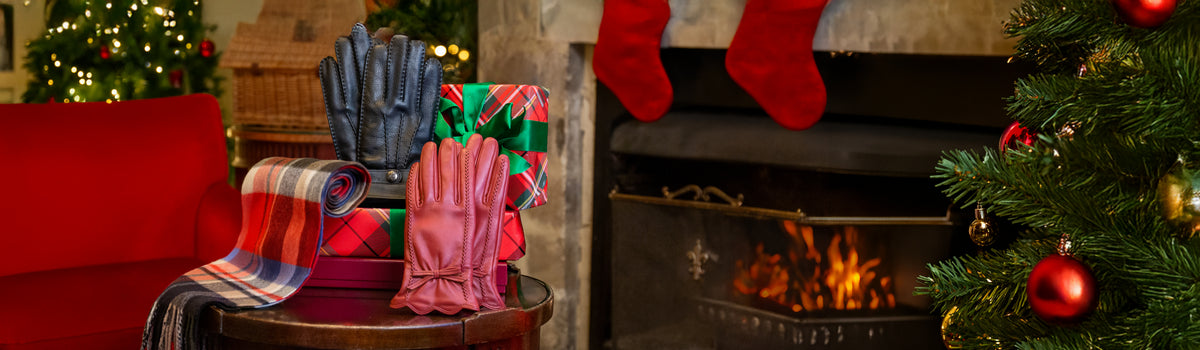 A cozy holiday scene with a round table displaying a pair of men's black leather gloves, a pair of women's red leather gloves, a cashmere plaid scarf, and a wrapped gift, set in front of a fireplace with red stockings and a Christmas tree.