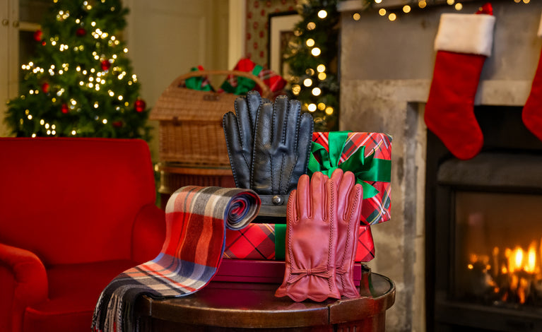 A coZy holiday scene with a round table displaying a pair of men's black leather gloves, a pair of women's red leather gloves, a cashmere plaid scarf, and a wrapped gift, set in front of a fireplace with red stockings and a Christmas tree.