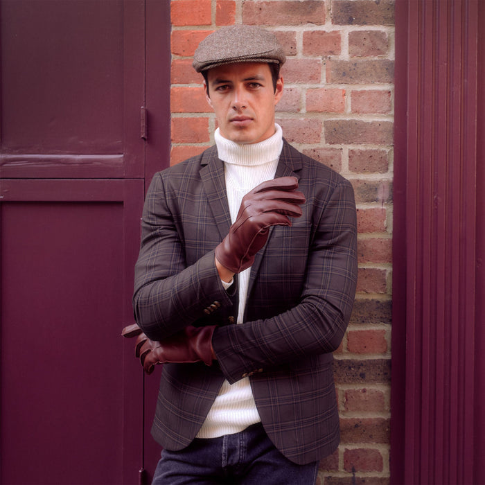 Man standing in front of a red brick wall wearing dark brown flat cap flecked with beige.
