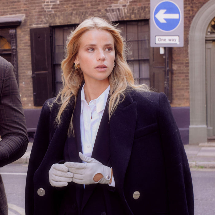 Woman wearing light grey driving gloves and a white shirt with black overcoat on a street.