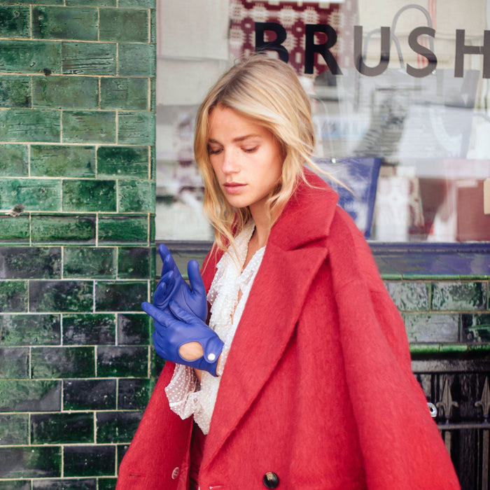 Woman standing by a green brick wall, wearing vibrant blue driving gloves with key hole opening.